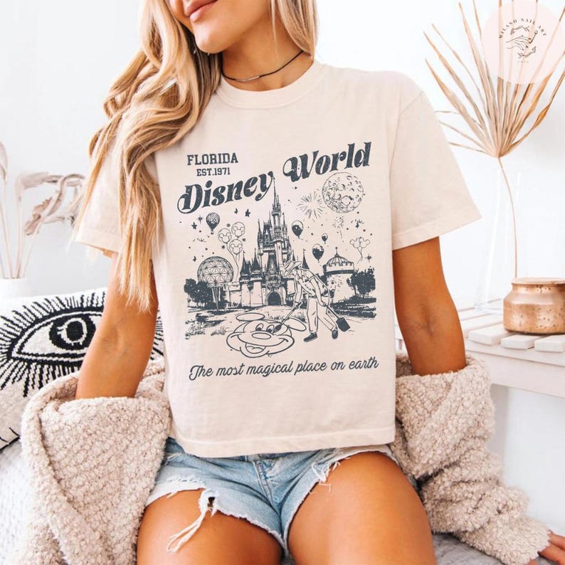 Może przedstawiać: Kremowa koszulka z grafiką Disney World. Grafika przedstawia zamek, Myszka Miki oraz tekst "Florida Est. 1971 Disney World" i "The most magical place on earth."