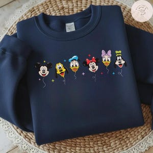 May include: Navy blue sweatshirt featuring embroidered Disney characters as balloons. Mickey Mouse, Pluto, Donald Duck, Minnie Mouse, Daisy Duck, and Goofy are depicted in vibrant colors. The design includes colorful star accents, creating a playful and whimsical aesthetic.