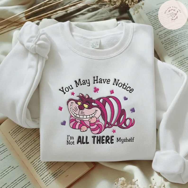 May include: White sweatshirt featuring a graphic of the Cheshire Cat from Alice in Wonderland. The cat is pink and purple with a wide grin. Text reads "You May Have Notice, I'm Not All There Myself."