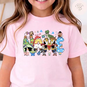以下が含まれることがあります： NAMEという文字が大きくプリントされたライトピンクのTシャツ。各文字はトイ・ストーリーのキャラクターにインスパイアされた模様や色で装飾されており、映画のキャストの小さなイラストが周囲に散りばめられています。