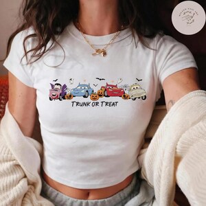 May include: A white short-sleeved t-shirt featuring a graphic of four cartoon cars dressed in Halloween costumes, including a vampire, a mummy, and a red race car, alongside orange pumpkins, spiderwebs, bats, and the text TRUNK OR TREAT.