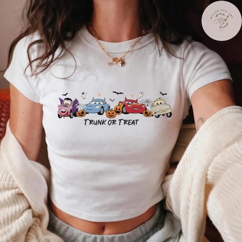 May include: A white short-sleeved t-shirt featuring a graphic of four cartoon cars dressed in Halloween costumes, including a vampire, a mummy, and a red race car, alongside orange pumpkins, spiderwebs, bats, and the text TRUNK OR TREAT.