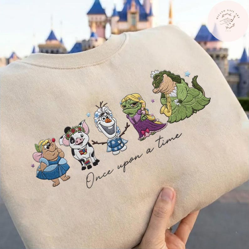 May include: Beige sweatshirt featuring embroidered cartoon characters, including a fairy, pig, snowman, and frog, with the text "Once upon a time." The characters are in various colors, and the sweatshirt has a crew neck.