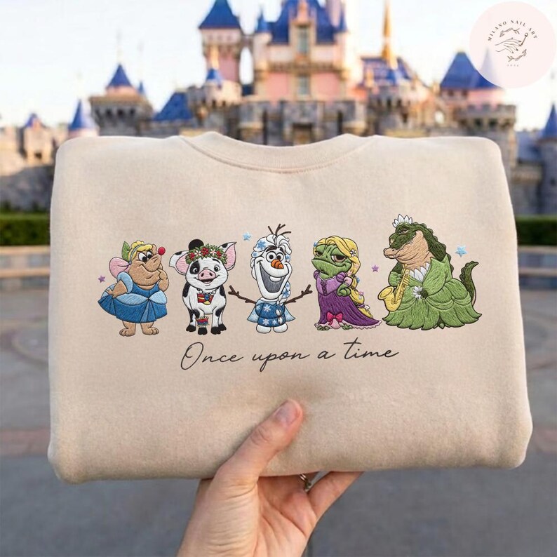 May include: A beige sweatshirt featuring embroidered cartoon characters. The design includes a fairy, pig, snowman, frog, and alligator. The phrase "Once upon a time" is written below the characters. The background shows a castle.