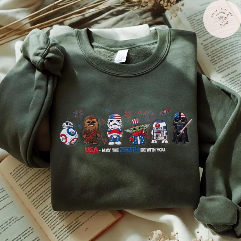 Op de afbeelding: Een olijfgroene sweatshirt met geborduurde Star Wars-personages in patriottische outfits. Het ontwerp bevat BB-8, Chewbacca, een Stormtrooper, Grogu, R2-D2 en Darth Vader, met de tekst "USA - MAY THE 250TH BE WITH YOU."