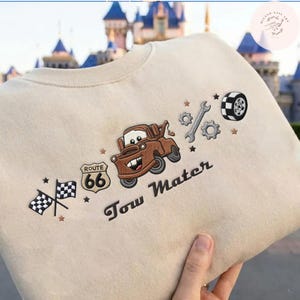 May include: A beige sweatshirt featuring embroidered designs including a brown tow truck, a Route 66 sign, two checkered racing flags, a wrench, gears, and a tire. The text Tow Mater is embroidered in dark brown script below the truck graphic.