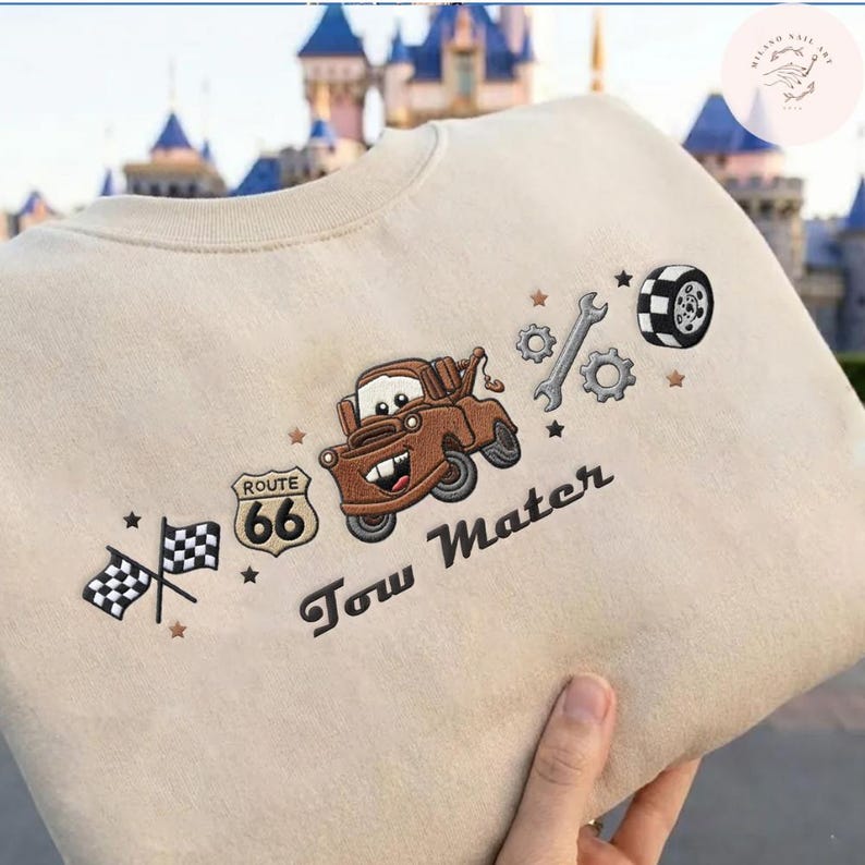 May include: A beige sweatshirt featuring embroidered designs including a brown tow truck, a Route 66 sign, two checkered racing flags, a wrench, gears, and a tire. The text Tow Mater is embroidered in dark brown script below the truck graphic.