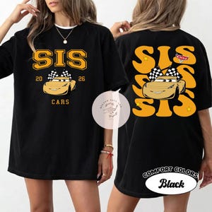 May include: Black t-shirt with a gold graphic featuring the word "SIS", the numbers "20" and "26", and a cartoon car with a checkered flag. The back of the shirt has a similar design. The shirt is labeled "Comfort Colors Black".