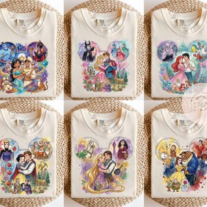 May include: A collection of six off-white t-shirts, each featuring a different watercolor-style graphic of Disney characters arranged in a Mickey Mouse ear silhouette. The designs include scenes from Aladdin, Sleeping Beauty, The Little Mermaid, Snow White, Tangled, and Beauty and the Beast.