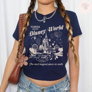 Może przedstawiać: Granatowa koszulka z nadrukiem zamku Kopciuszka, Myszki Miki i fajerwerk&oacute;w, z napisem "FLORIDA EST.1971 Disney World" i "The most magical place on earth". Koszulka jest połączona z dżinsowymi szortami i brązową torebką.