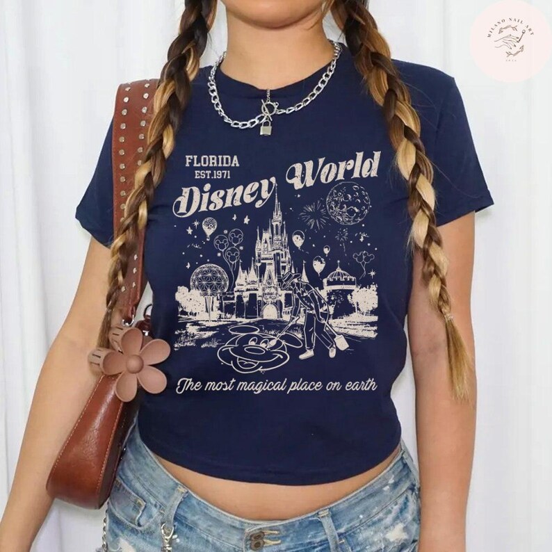 Może przedstawiać: Granatowa koszulka z nadrukiem zamku Kopciuszka, Myszki Miki i fajerwerk&oacute;w, z napisem "FLORIDA EST.1971 Disney World" i "The most magical place on earth". Koszulka jest połączona z dżinsowymi szortami i brązową torebką.