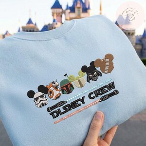 May include: Pale blue sweatshirt with embroidered Disney and Star Wars character designs. Includes Mickey Mouse ears, BB-8, Boba Fett, Darth Vader, and Chewbacca. The text "DISNEY CREW" is embroidered on the front.