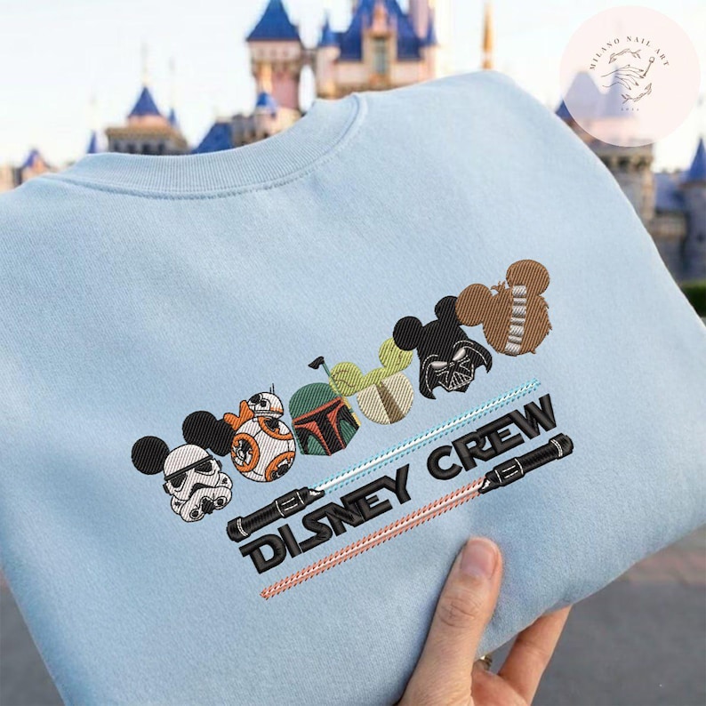 May include: Pale blue sweatshirt with embroidered Disney and Star Wars character designs. Includes Mickey Mouse ears, BB-8, Boba Fett, Darth Vader, and Chewbacca. The text "DISNEY CREW" is embroidered on the front.