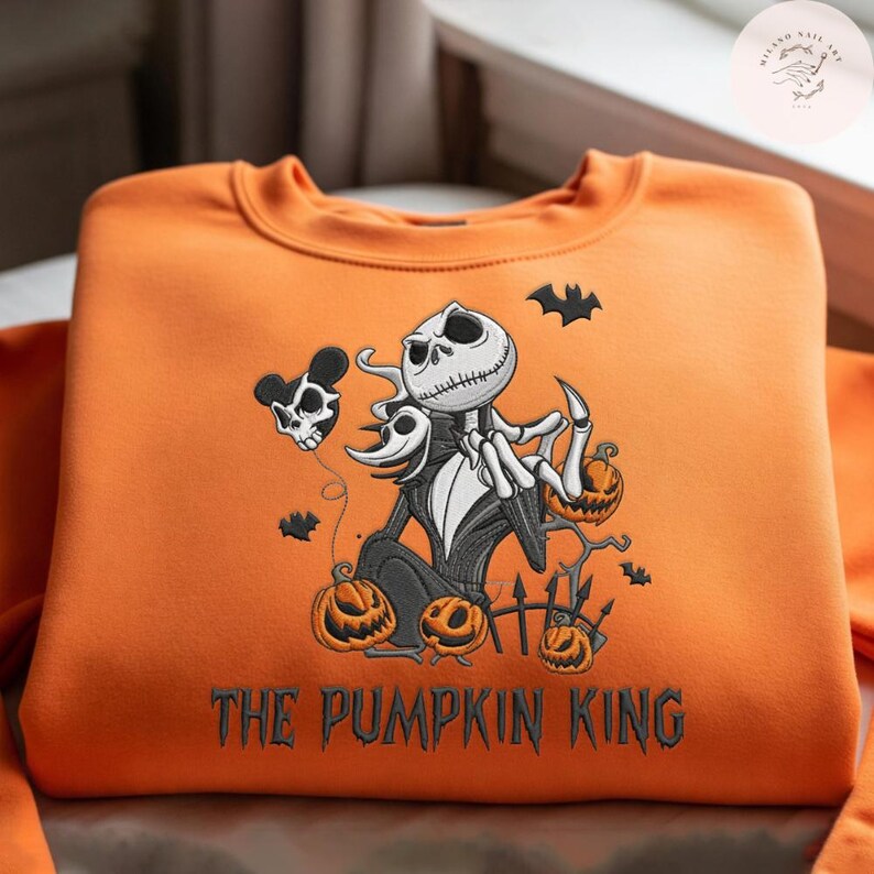 May include: An orange crewneck sweatshirt featuring an embroidered graphic of Jack Skellington, a ghost dog, and several jack-o'-lanterns. The design includes a skull-shaped balloon, small bats, and the text THE PUMPKIN KING in black lettering.
