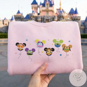 May include: A light pink sweatshirt featuring embroidered Mickey Mouse-shaped balloons. Each balloon is designed with a different character theme, including a cowboy, alien, and dog. Blue stars are scattered around the balloons.