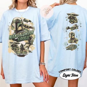 May include: Light blue t-shirt with Star Wars: The Mandalorian and Grogu graphics. The front shows the Mandalorian helmet and Grogu, with the text "THIS IS THE WAY." The back features character and ship illustrations. The shirt is labelled "Comfort Colors Light Blue."