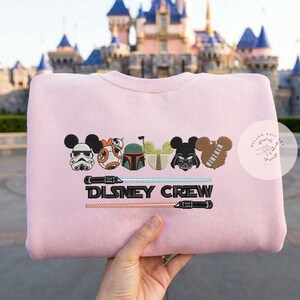 May include: A pale pink sweatshirt with embroidered Disney and Star Wars characters, including Mickey Mouse, BB-8, and Darth Vader. The text "DISNEY CREW" is embroidered below the characters, with lightsabers. A fun, themed apparel item.