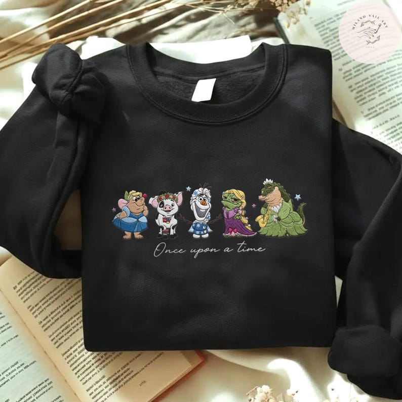 May include: Black sweatshirt featuring an embroidered design of cartoon characters. The design includes a fairy, pig, snowman, princess, and alligator. The phrase "Once upon a time" is written below the characters in a cursive font.