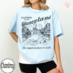 May include: Light blue t-shirt with a Disneyland graphic. The graphic features a street scene with a castle, fireworks, and Mickey Mouse. Text includes "CALIFORNIA EST.1955", "Disneyland", and "The happiest place on earth."
