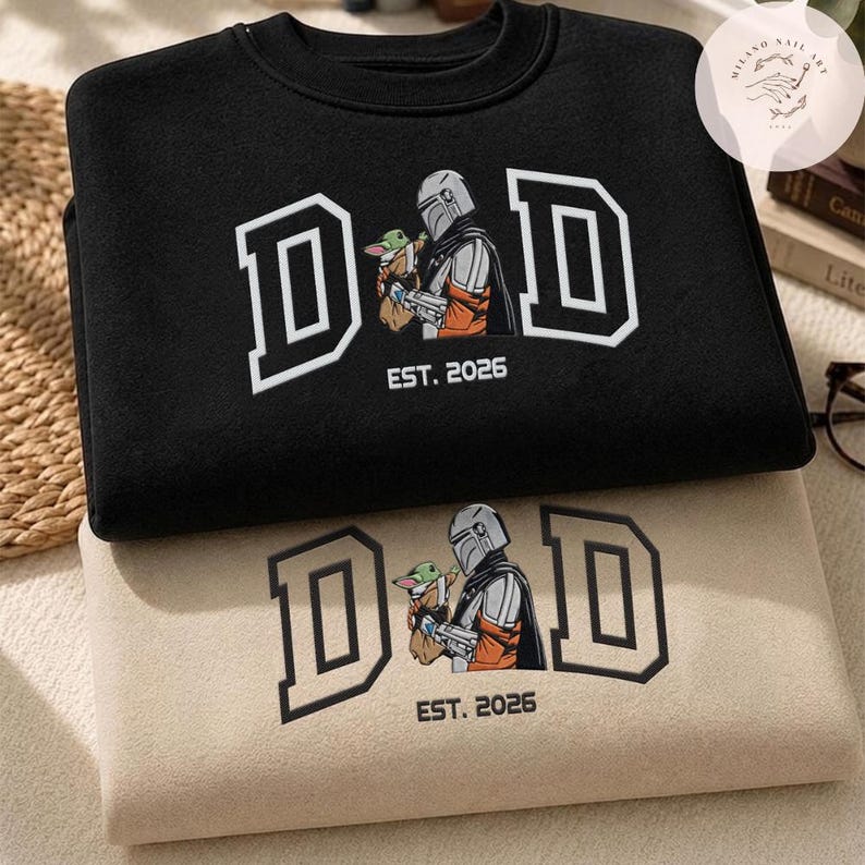 May include: Two sweatshirts, one black and one beige, are stacked. Both feature the word "DAD" in a white outlined font, with an image of a Mandalorian character holding a small green creature. The text "EST. 2026" is below the image.