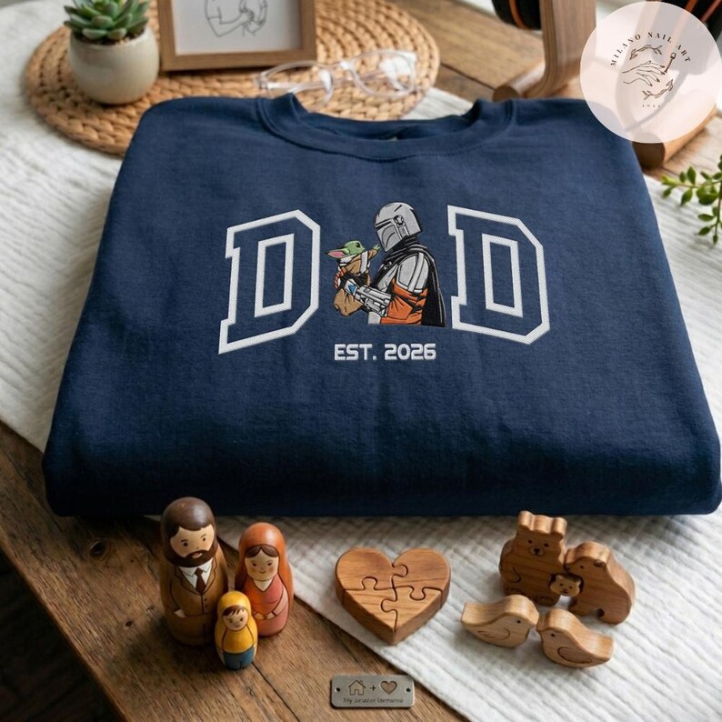 May include: Navy blue sweatshirt with the word "DAD" in white block letters, featuring a graphic of a Mandalorian character and Grogu. The text "EST. 2026" is below the graphic. Wooden family figurines and a wooden heart puzzle are in the foreground.