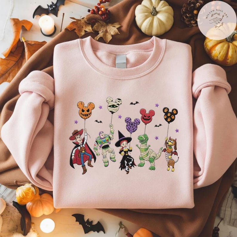 May include: A light pink crewneck sweatshirt featuring a graphic print of five cartoon characters dressed in Halloween costumes, each holding a Mickey Mouse-shaped balloon. The characters include a cowboy, a space ranger, a cowgirl, a dinosaur, and a dog.