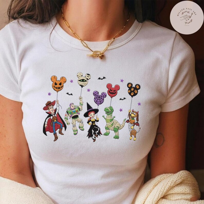 May include: A white short-sleeved t-shirt featuring a graphic print of various cartoon characters dressed in Halloween costumes, each holding a Mickey Mouse-shaped balloon. The design includes small bats and purple stars scattered around the characters.