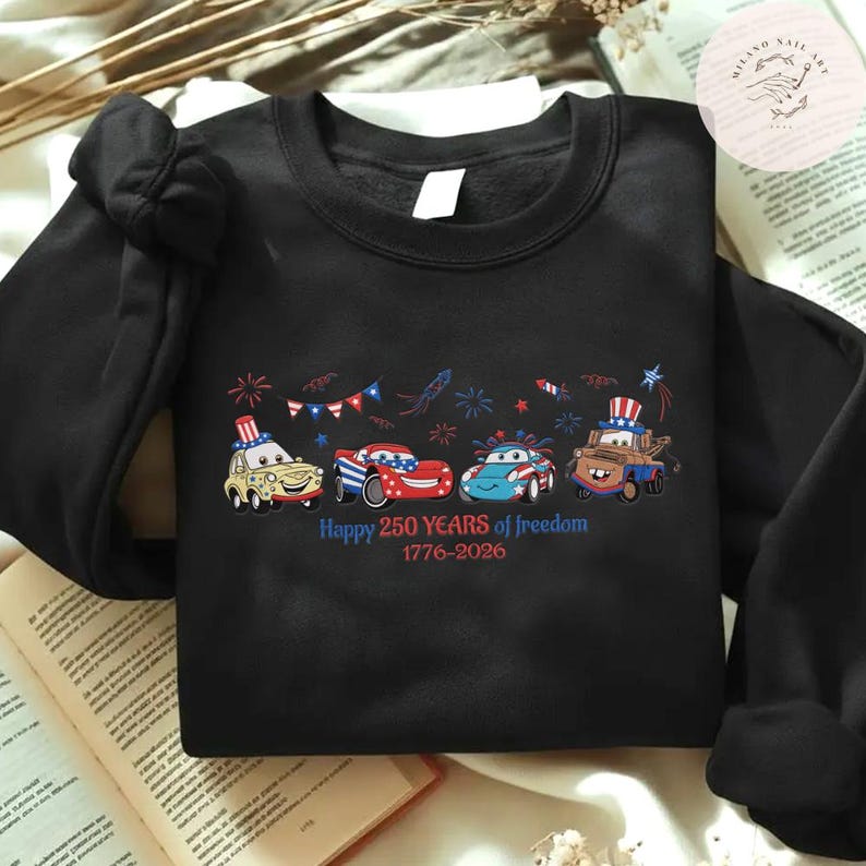 Pode incluir: Camisola preta com um gr&aacute;fico de carros de desenho animado em vermelho, branco e azul, celebrando o 250&ordm; anivers&aacute;rio da liberdade. O texto "Happy 250 Years of Freedom 1776-2026" est&aacute; impresso abaixo dos carros.