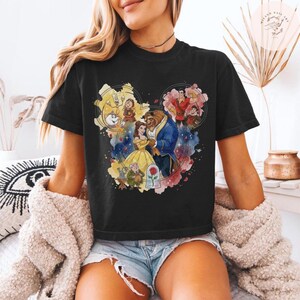 May include: A black short-sleeve t-shirt featuring a colorful watercolor-style graphic of characters from Beauty and the Beast arranged in a Mickey Mouse head silhouette, including Belle, the Beast, Lumiere, Cogsworth, Mrs. Potts, and a rose.