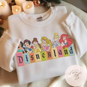 May include: White sweatshirt with an embroidered design of Disney princesses and the word "Disneyland". The design features Jasmine, Snow White, Belle, Aurora, Cinderella, and Ariel. The sweatshirt has a crew neck and long sleeves.