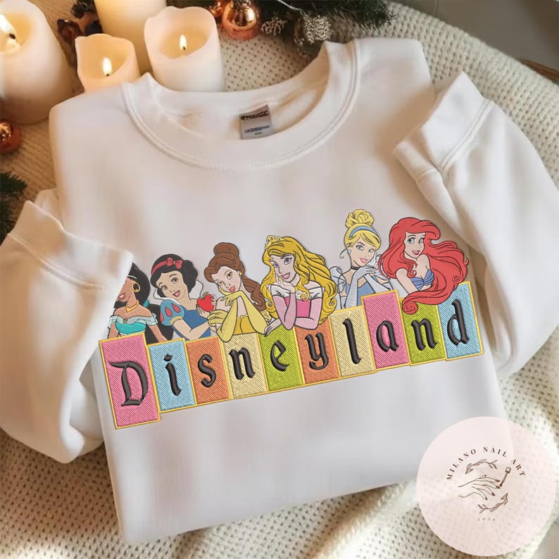 May include: White sweatshirt with an embroidered design of Disney princesses and the word "Disneyland". The design features Jasmine, Snow White, Belle, Aurora, Cinderella, and Ariel. The sweatshirt has a crew neck and long sleeves.