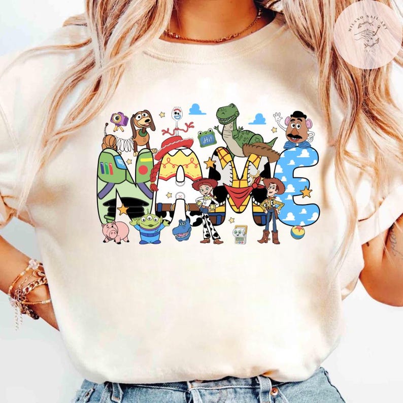 以下が含まれることがあります： クリーム色のTシャツで、大きなブロック体で「NAME」という文字がカラフルにプリントされています。文字には、ウッディ、バズ・ライトイヤー、ジェシー、レックス、ミスター・ポテトヘッドなど、トイ・ストーリーのキャラクターたちが描かれています。