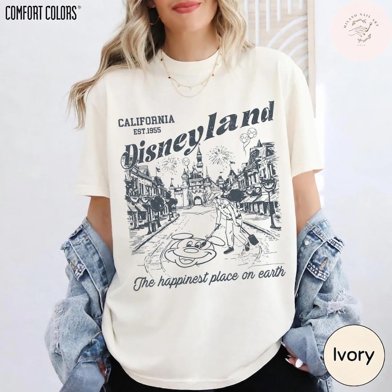 May include: Ivory-colored t-shirt with a Disneyland graphic. The graphic features a street scene with a castle, fireworks, and Mickey Mouse. The text reads "California Disneyland Est.1955" and "The happiest place on earth."