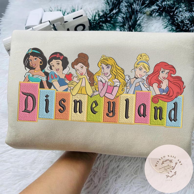May include: A beige sweatshirt with an embroidered design of Disney princesses above the word "Disneyland" in colourful, block letters. The princesses include Jasmine, Snow White, Belle, Aurora, Cinderella, and Ariel.