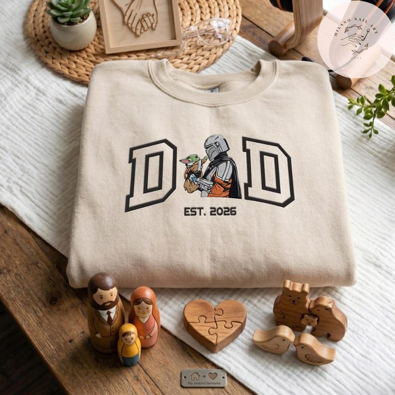 May include: Beige sweatshirt with the word "DAD" in black block letters, featuring a graphic of a Mandalorian character and Grogu. The text "EST. 2026" is below the graphic. Wooden family figurines and a heart-shaped puzzle are in the foreground.