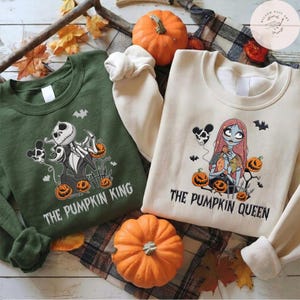 May include: A dark green sweatshirt featuring a graphic of a skeleton character and the text THE PUMPKIN KING, alongside a cream sweatshirt featuring a graphic of a female skeleton character and the text THE PUMPKIN QUEEN, both with pumpkins.