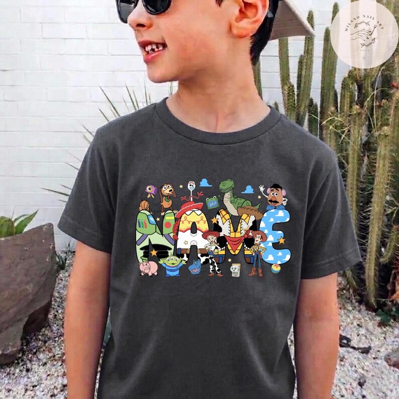 以下が含まれることがあります： ダークグレーの半袖Tシャツ。中央には「NAME」という文字が大きくプリントされており、各文字の中にはウッディ、バズ・ライトイヤー、レックス、ミスター・ポテトヘッドなど、トイ・ストーリーの様々なキャラクターのイラストが鮮やかな色で描かれています。