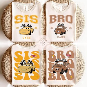 May include: Four white t-shirts with cartoon car graphics. Two shirts say "SIS" with a yellow car and checkered flags, and the other two say "BRO" with a brown truck and checkered flags. The shirts are on a woven background.