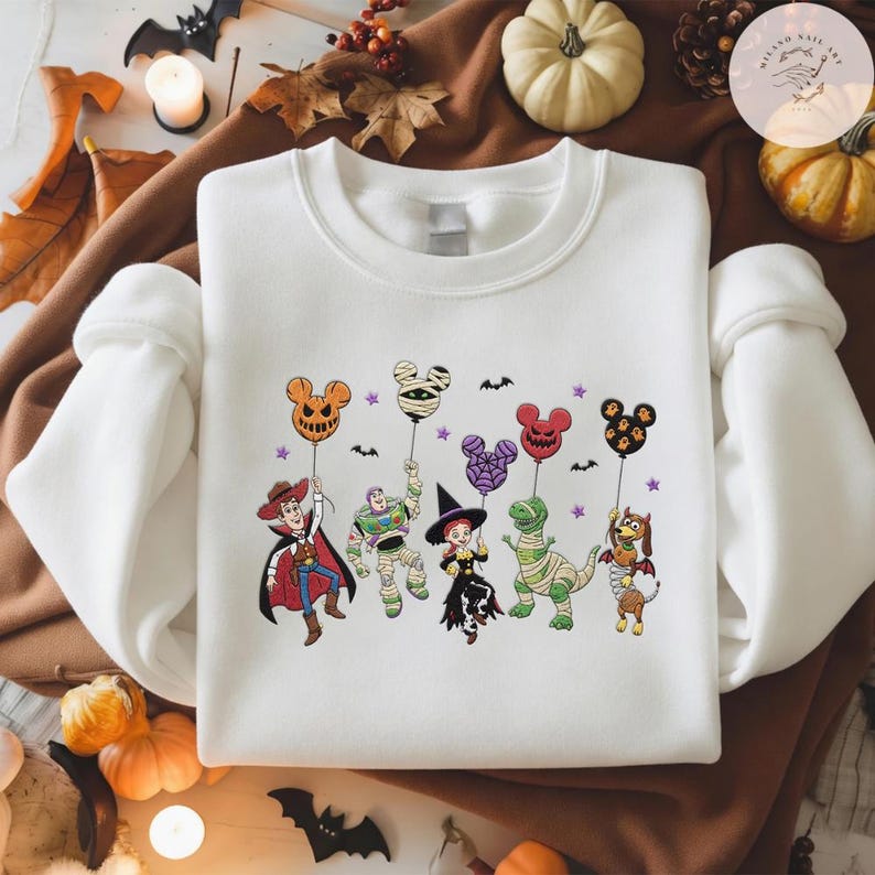 May include: A white crewneck sweatshirt featuring a graphic print of five cartoon characters dressed in Halloween costumes, each holding a Mickey Mouse-shaped balloon. The characters include a cowboy, a space ranger, a cowgirl, a dinosaur, and a dog.