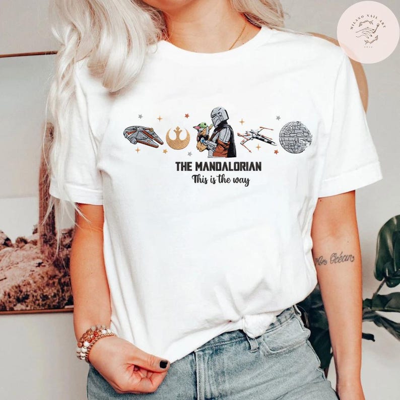 May include: White t-shirt featuring a graphic design from The Mandalorian. The design includes illustrations of the Millennium Falcon, a symbol, the Mandalorian with Grogu, an X-wing, and the Death Star. Text reads "THE MANDALORIAN" and "This is the way."