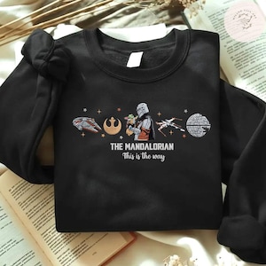 May include: Black sweatshirt featuring a graphic from "The Mandalorian." The design includes the text "THE MANDALORIAN This is the way" along with illustrations of a spaceship, a symbol, the Mandalorian with Grogu, an X-wing, and the Death Star.