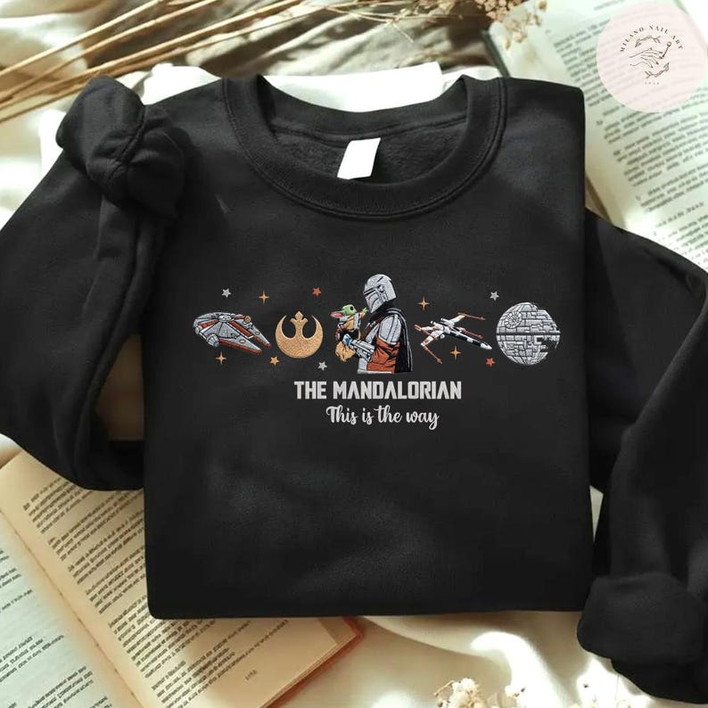 May include: Black sweatshirt featuring a graphic from "The Mandalorian." The design includes the text "THE MANDALORIAN This is the way" along with illustrations of a spaceship, a symbol, the Mandalorian with Grogu, an X-wing, and the Death Star.