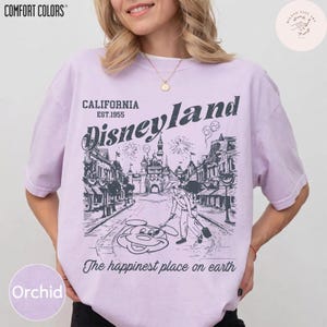 May include: Orchid-colored t-shirt with a Disneyland graphic. The graphic features the Disneyland logo, a street scene, and Mickey Mouse. Text on the shirt reads "CALIFORNIA EST.1955 Disneyland" and "The happiest place on earth."