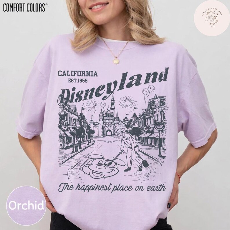 May include: Orchid-colored t-shirt with a Disneyland graphic. The graphic features the Disneyland logo, a street scene, and Mickey Mouse. Text on the shirt reads "CALIFORNIA EST.1955 Disneyland" and "The happiest place on earth."