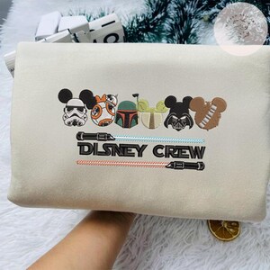 May include: A beige embroidered pillowcase with Disney characters in a Mickey Mouse style. The design includes a Stormtrooper, BB-8, Boba Fett, Yoda, Darth Vader, and Chewbacca. The text "DISNEY CREW" is embroidered below the characters, with lightsabers.