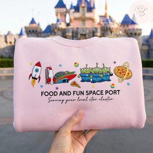 May include: A light pink sweatshirt with a colorful graphic featuring a rocket, aliens, pizza, and the text "FOOD AND FUN SPACE PORT". The sweatshirt has the phrase "Serving your local star cluster" below the graphic.