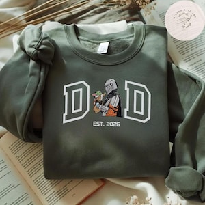 May include: Olive green sweatshirt with the word "DAD" in white block letters. The graphic features a Mandalorian character holding Grogu, with "EST. 2026" below. The sweatshirt is folded on a surface with a book.