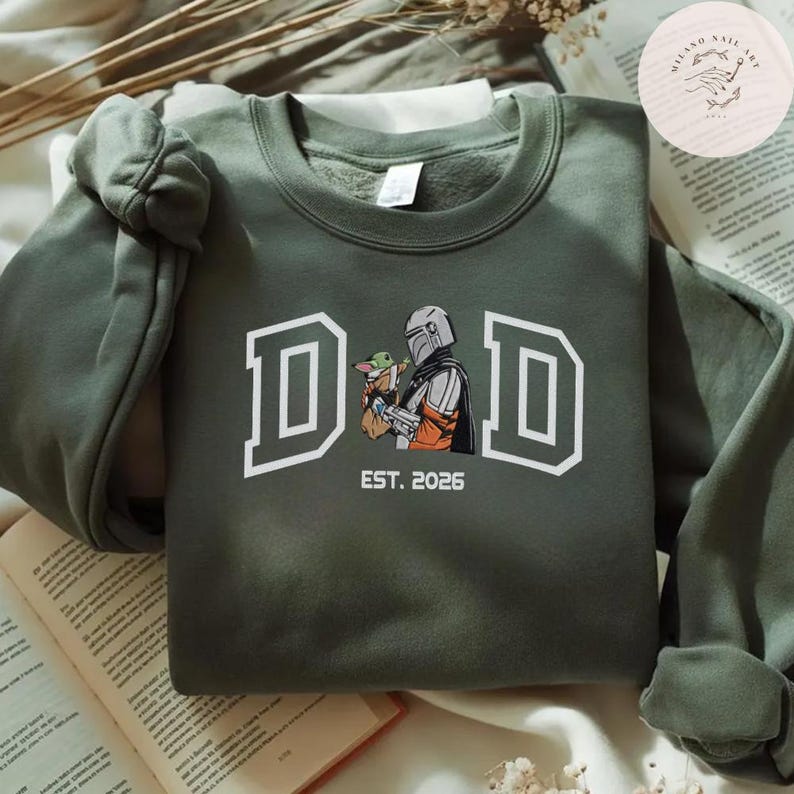 May include: Olive green sweatshirt with the word "DAD" in white block letters. The graphic features a Mandalorian character holding Grogu, with "EST. 2026" below. The sweatshirt is folded on a surface with a book.