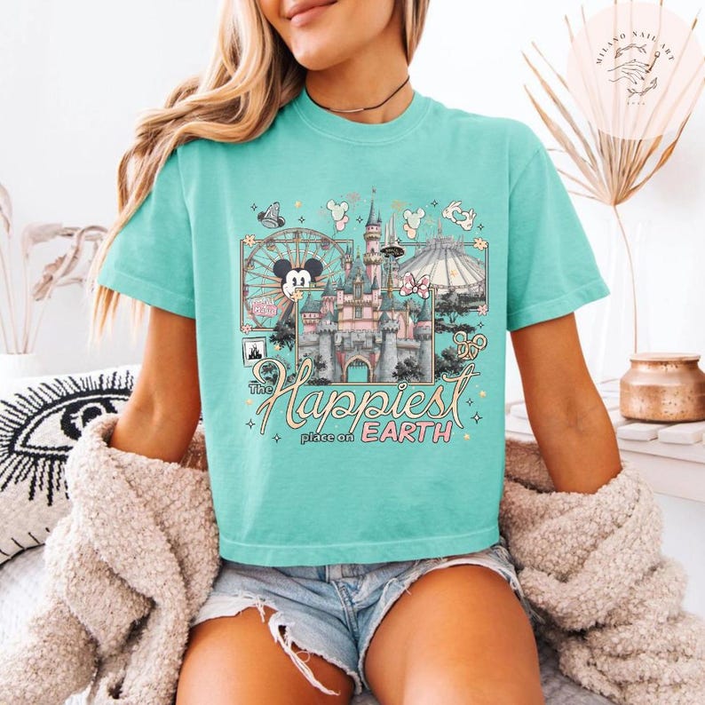 May include: A turquoise t-shirt featuring a colorful graphic of a castle, Ferris wheel, and other theme park elements. The text "The Happiest Place on Earth" is displayed below the graphic. The shirt has short sleeves and a crew neck.