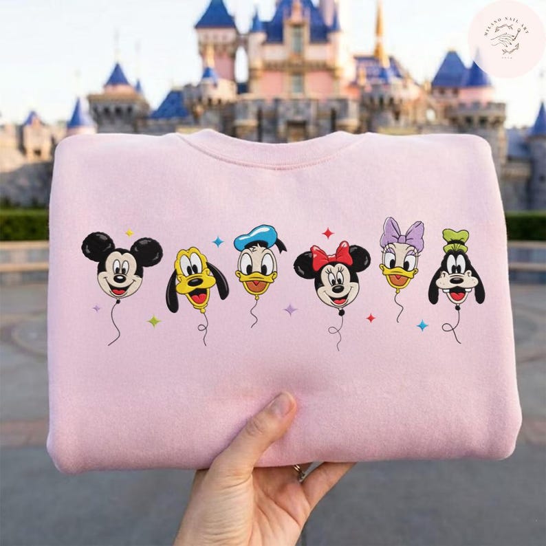 May include: A light pink sweatshirt featuring a graphic of Mickey Mouse, Pluto, Donald Duck, Minnie Mouse, Daisy Duck, and Goofy as balloon characters. Each character is depicted with a smiling face and attached to a string. The background shows a castle.